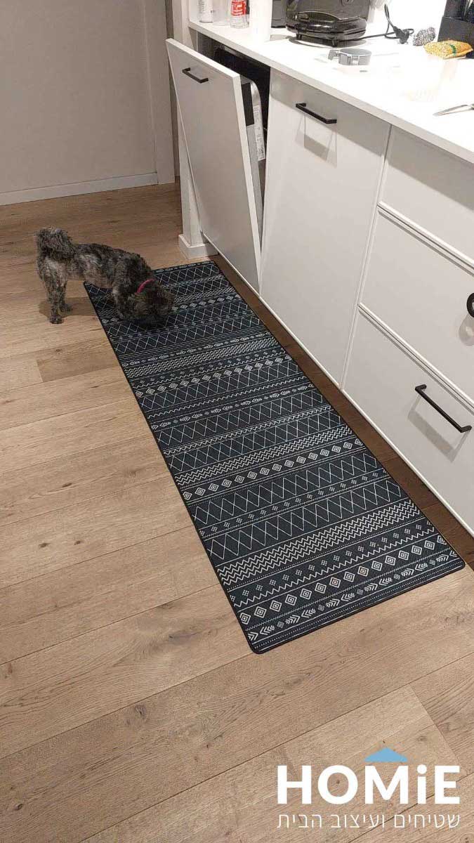 שטיח גומי ארוך בצבע שחור-לבן עם דוגמה גיאומטרית, מונח על רצפת עץ במטבח, ולצידו כלב קטן.